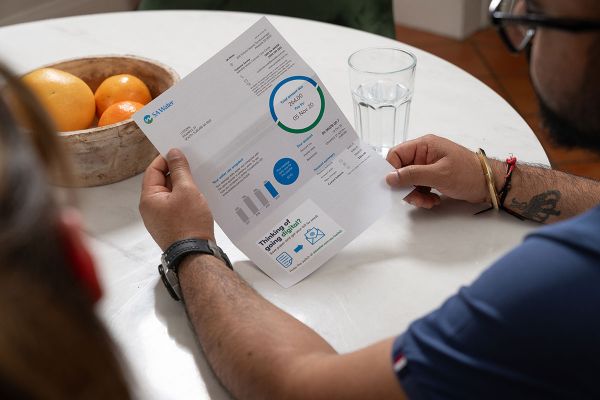 Man sitting at a table holding an SA Water bill, with a glass of water and a bowl of fruit nearby.